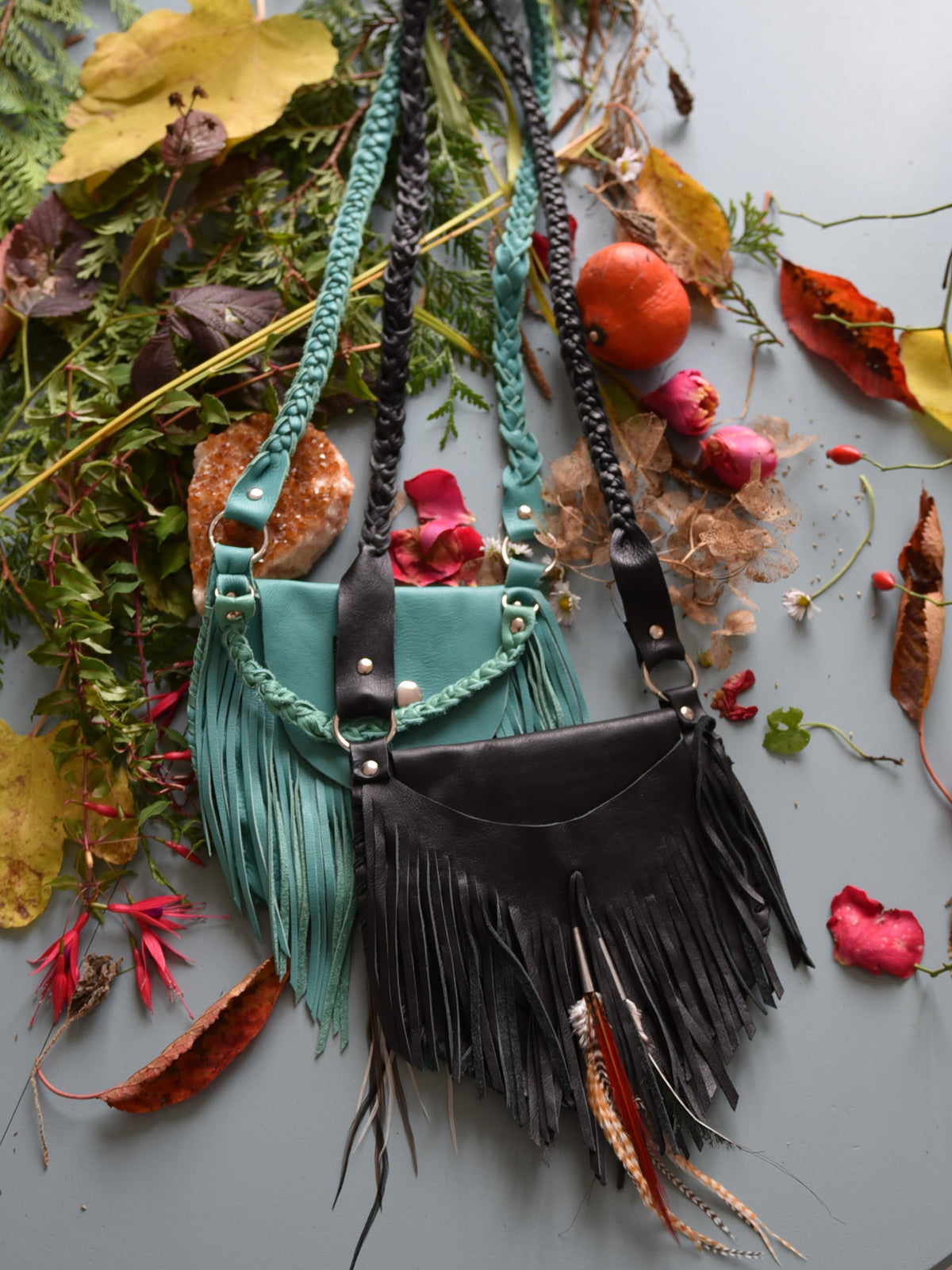 Ruby Feathers Achack leather fringe bags in teal and black with braided straps, displayed on a rustic autumn backdrop.