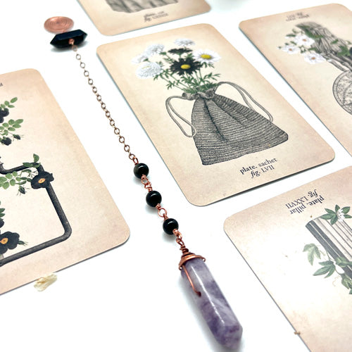 Ruby Feathers pendulum featuring a purple amethyst point, black beads, and copper wire on a background of botanical cards.