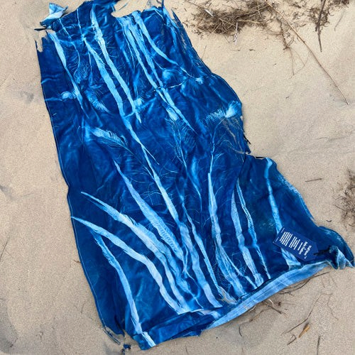 Ruby Feathers upcycled cotton t-shirt with blue sun-printed feather and lace patterns lying on sandy beach
